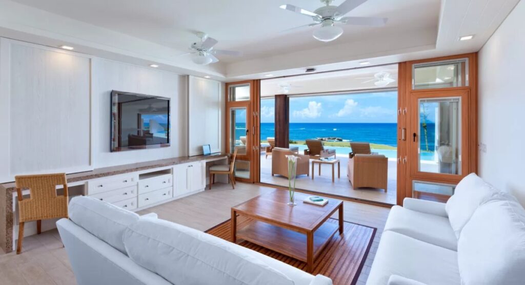 Luxurious beachfront living room with open sliding doors leading to a patio overlooking the ocean, featuring white sofas and wooden furniture