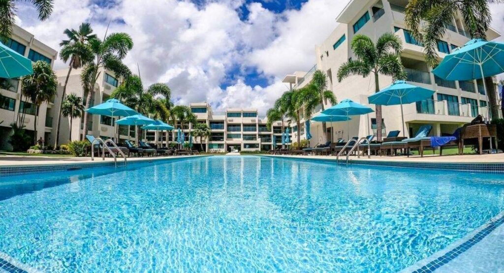 Inviting resort pool with crystal clear blue water, surrounded by white lounge chairs under turquoise umbrellas, tropical palm trees, and a view of a modern resort building under a sunny sky.