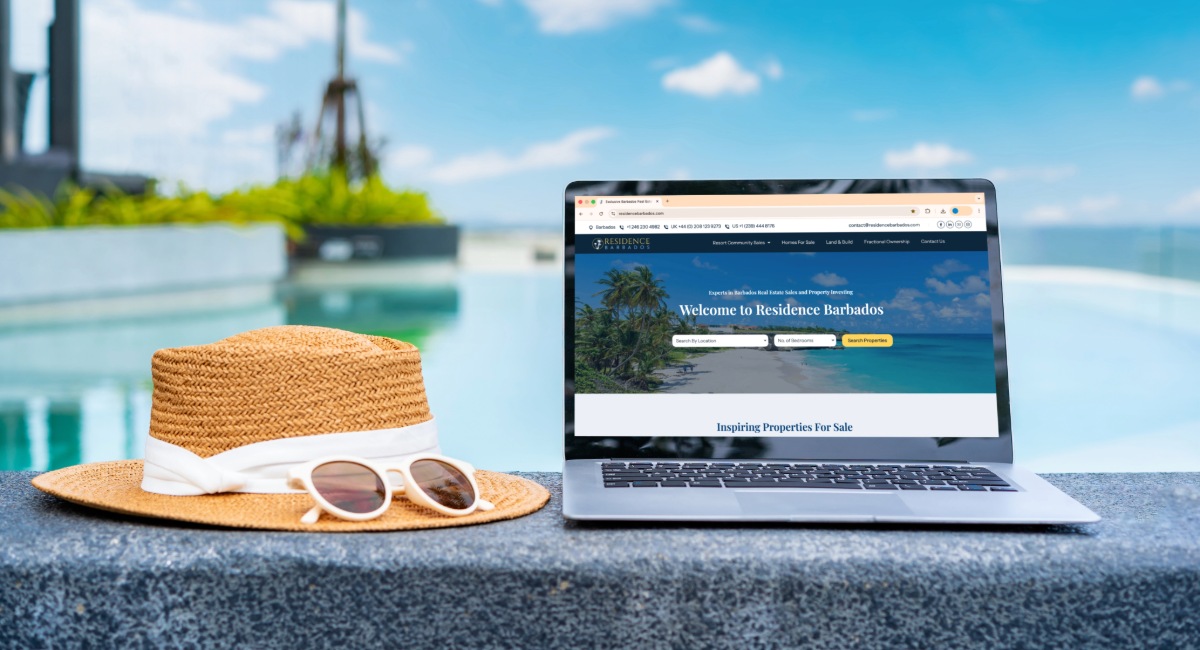 Digital nomad laptop setup displaying Residence Barbados website on outdoor table with straw hat and sunglasses overlooking Caribbean harbor - remote work paradise in Barbados