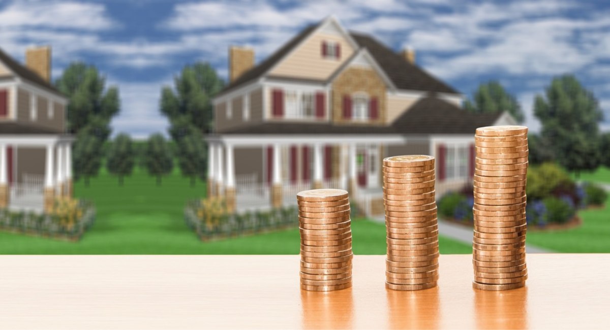 Three stacks of coins increasing in height, positioned in the foreground with a blurred two-story house and suburban lawn, showcasing taxes and costs on Barbados properties